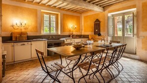A rustic kitchen with wooden beams, a large dining table, and a view of greenery outside.