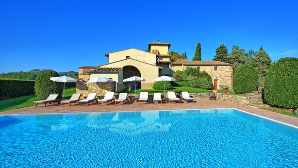 Ein Poolbereich mit Liegestühlen und Sonnenschirmen, ein Steinhaus und ein klarer, blauer Himmel.