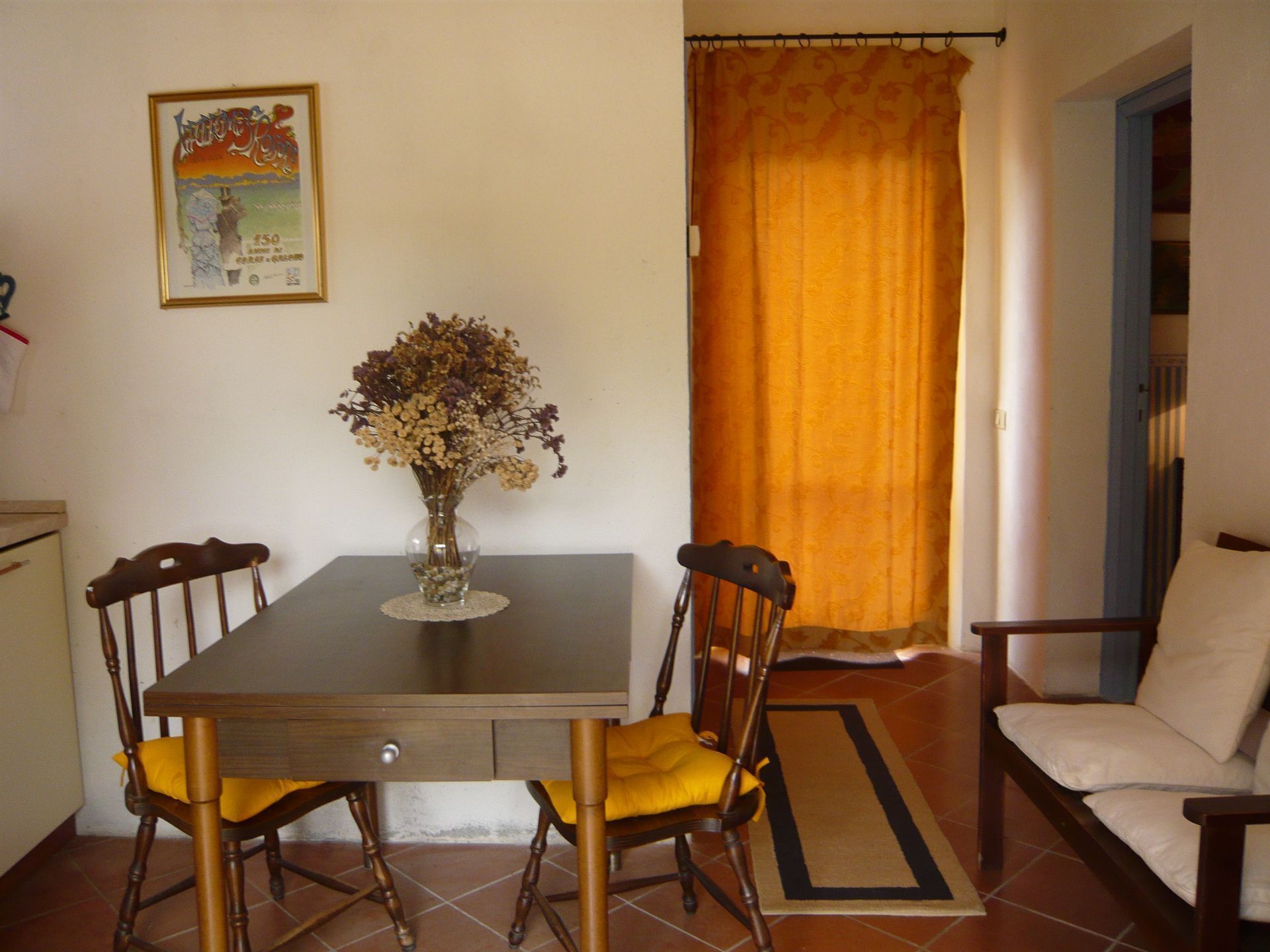 A dining area with a table, chairs, a vase with flowers, a framed picture on the wall, and a curtain.