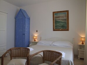 A bedroom with a bed, two wicker chairs, a blue door, a painting, and two bedside lamps.