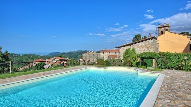 Une piscine aux eaux d’un bleu limpide, dont le fond est recouvert de carreaux de pierre, entourée d’un mur de pierre et d’une clôture en bois, et qui offre une vue panoramique sur un village et des montagnes.