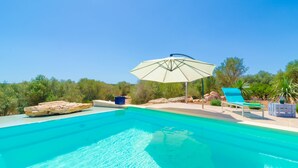 A clear blue pool with a lounge chair and umbrella, surrounded by greenery and rocks.