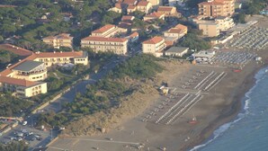 Vista aérea de un pueblo costero con playa, hileras de sombrillas y edificios de techos rojos.