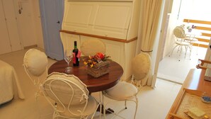 Un comedor con una mesa redonda, sillas blancas, una botella de vino y una cesta de fruta.