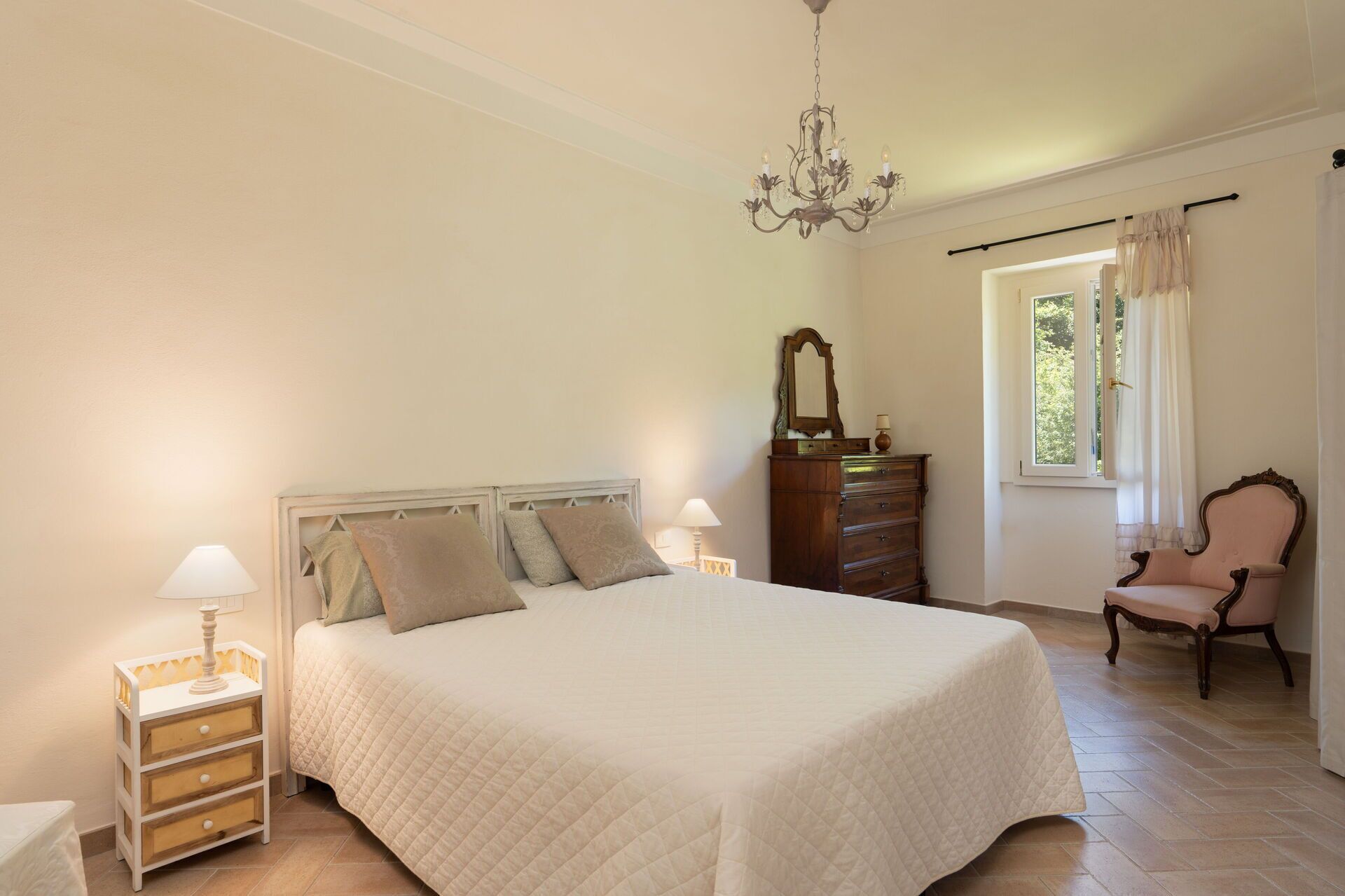 Une chambre à coucher avec un grand lit, des tables de chevet avec des lampes, une commode, une chaise et une fenêtre avec des rideaux.