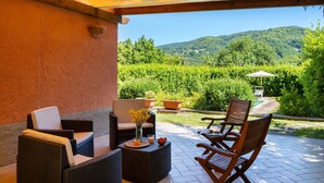 Un patio cubierto con mobiliario de mimbre, una mesa con un jarrón de flores y vistas a un jardín exuberante y colinas al fondo.