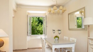Un comedor con una mesa y sillas blancas, una araña, una ventana con vistas a la vegetación y un espejo en la pared.