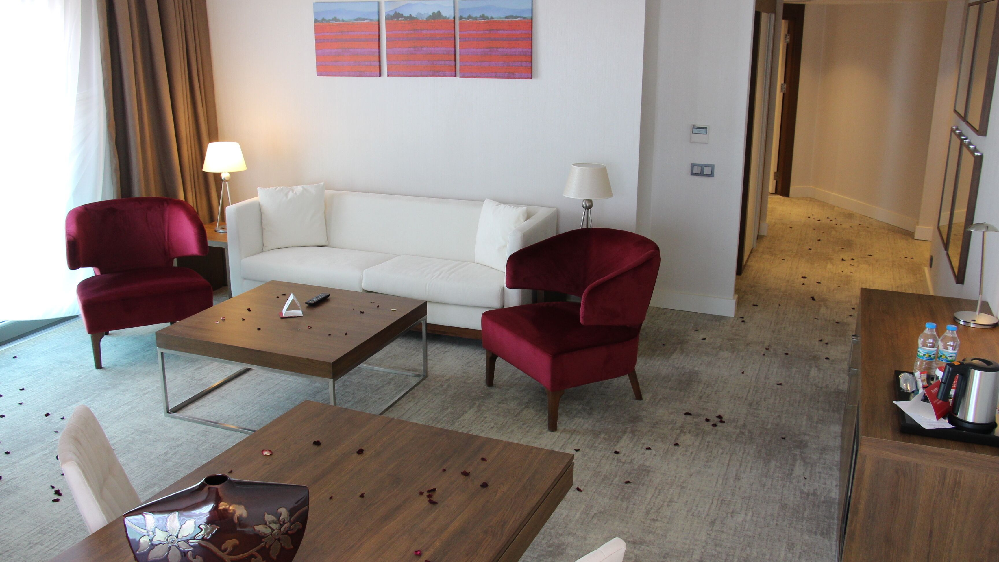 A hotel room with a white sofa, two red armchairs, a wooden coffee table, and a small table with a bottle and a kettle.