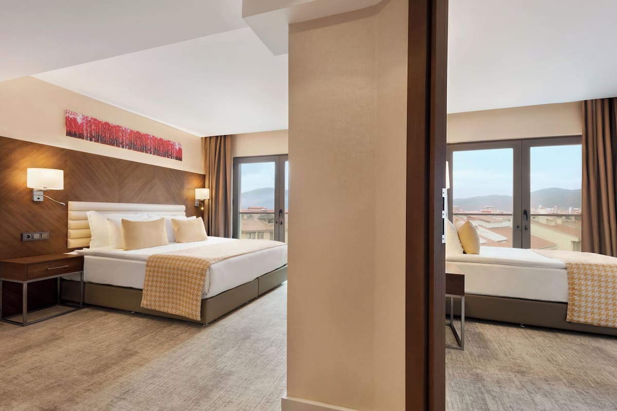 A hotel room with a large bed, a desk, a chair, a nightstand, a wall-mounted lamp, and a window with a view of the cityscape.