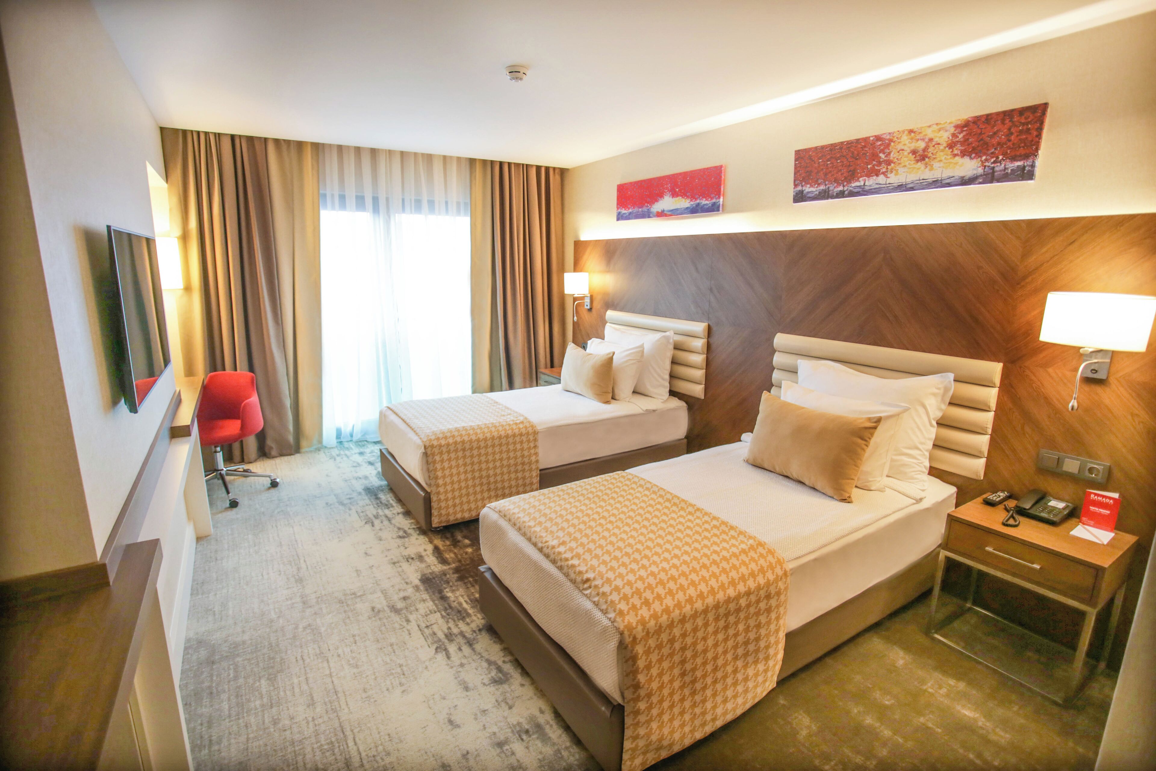 A hotel room with two beds, a desk, a chair, and a nightstand with a phone and a red sign.