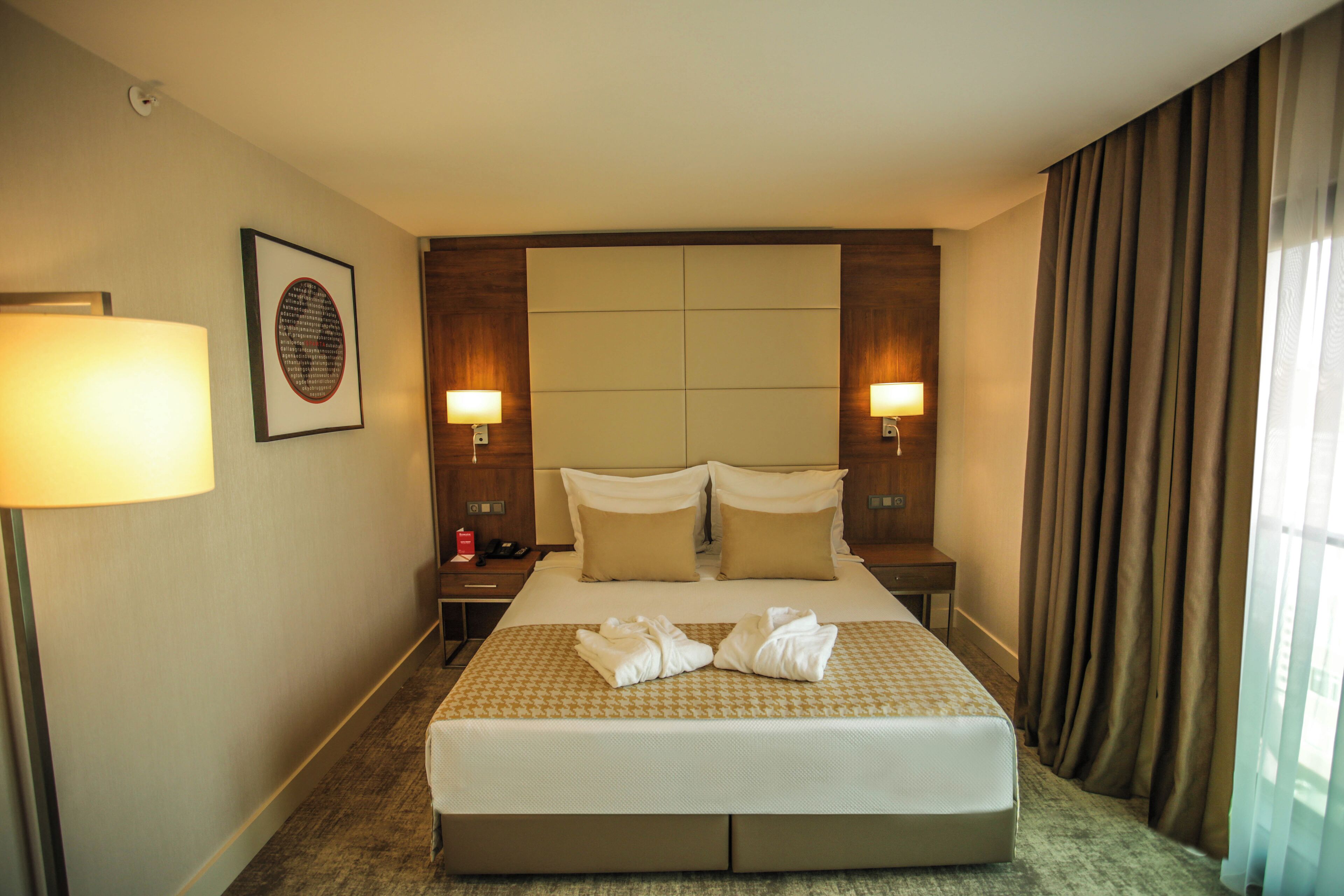 A hotel room with a large bed, two bedside lamps, a headboard, a framed picture, and a window with curtains.