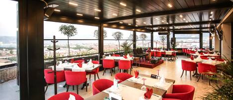 A restaurant with red chairs and tables, large windows, and a view of the city.