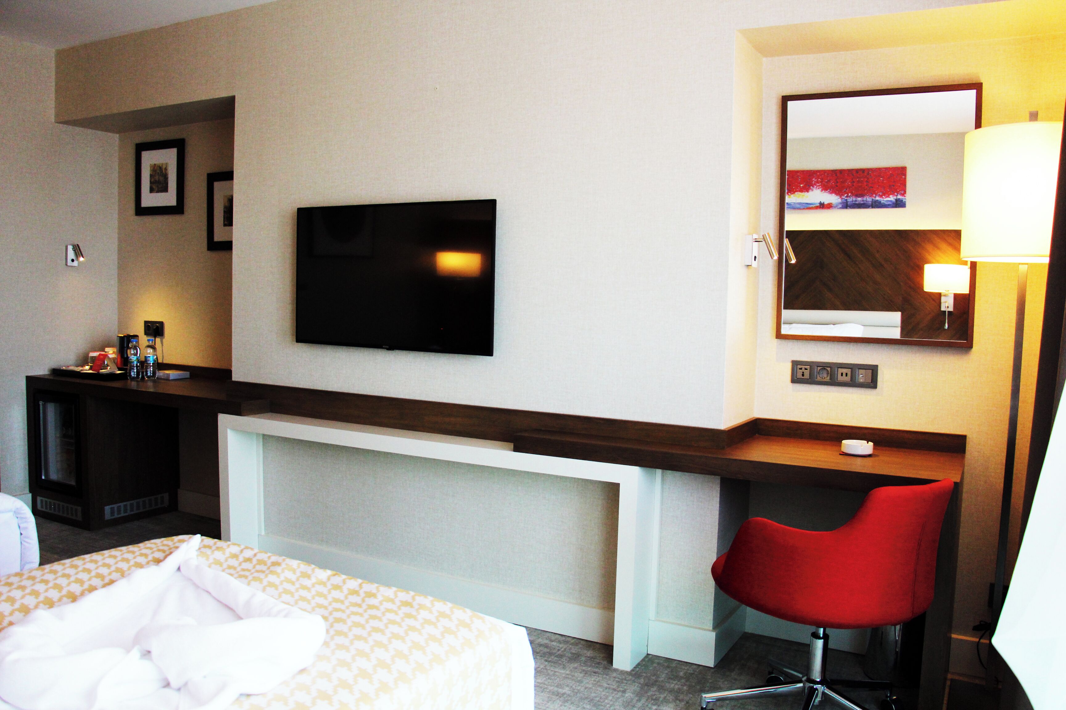 Una habitación de hotel con cama, escritorio, televisor, silla y espejo.