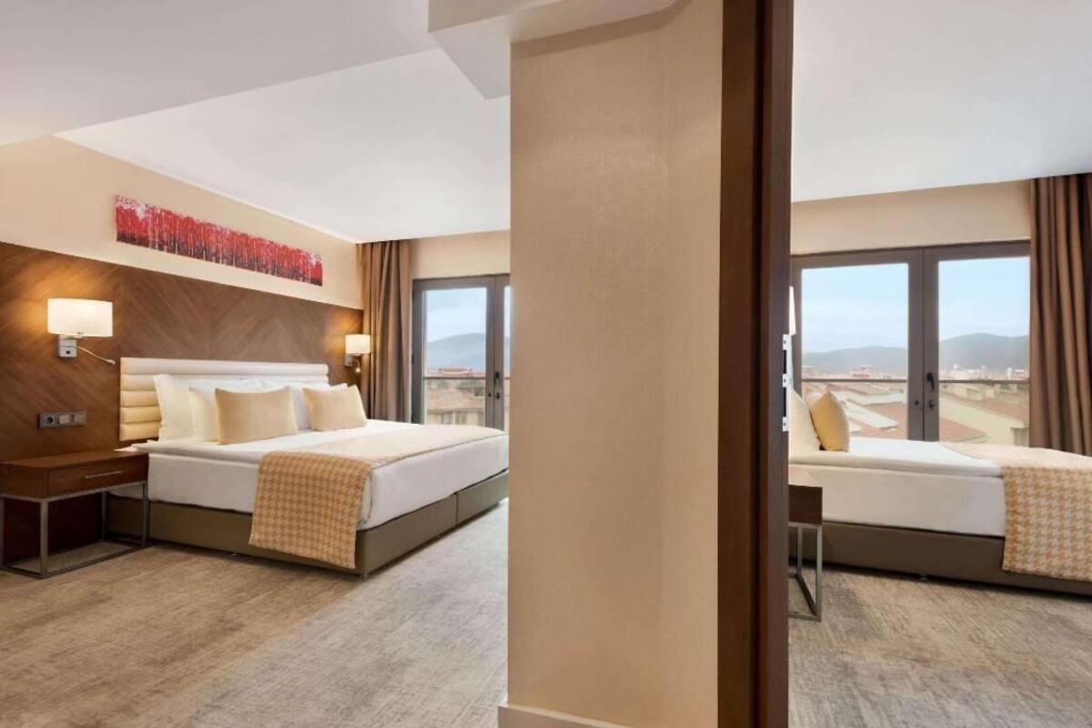A hotel room with a large bed, bedside tables, a wall-mounted lamp, and a large window offering a city view.