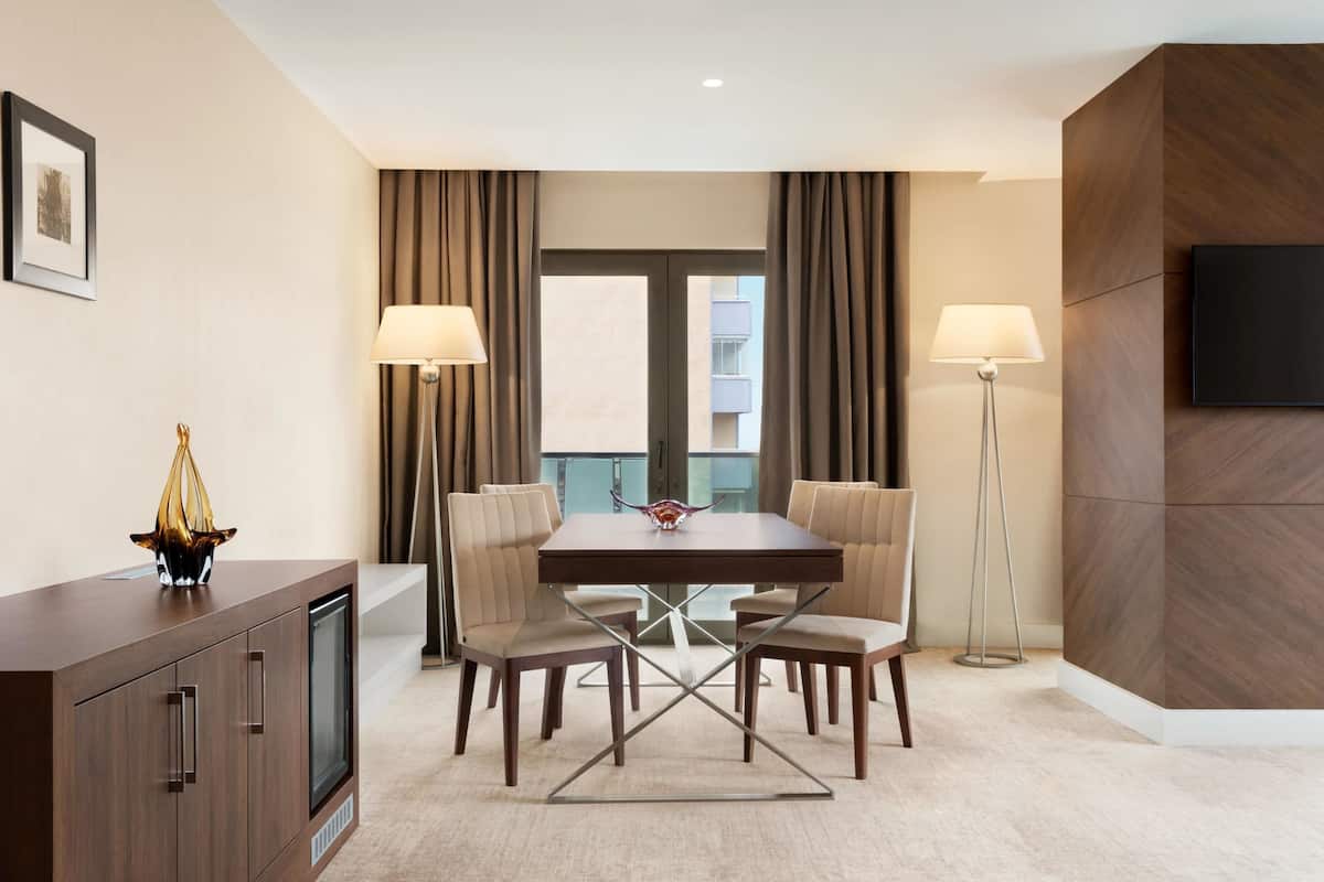 A modern hotel room with a dining table, two chairs, a TV mounted on the wall, and a floor lamp.
