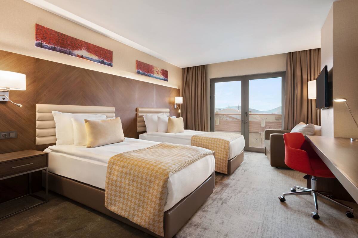 A hotel room with two beds, a desk, a chair, and a large window.
