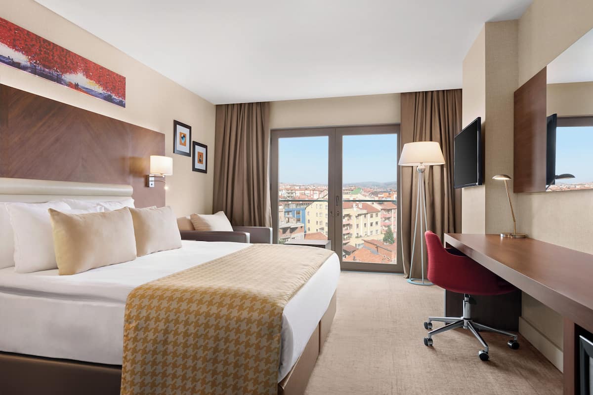 A hotel room with a large bed, a desk, a chair, a TV, and a window with a city view.