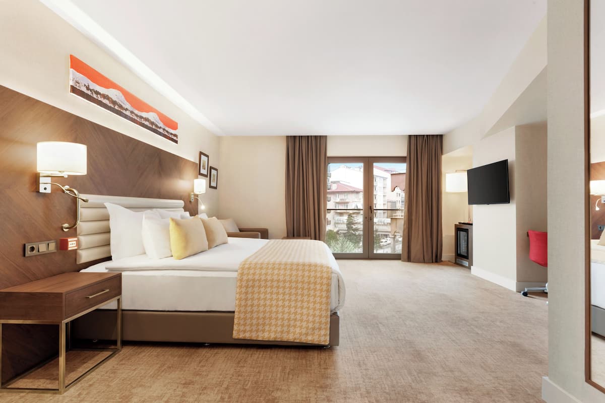 A hotel room with a bed, bedside table, lamp, artwork, television, and a view of buildings through a window.