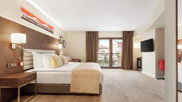 A hotel room with a bed, bedside table, lamp, artwork, television, and a view of buildings through a window.