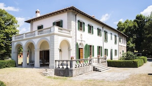 A two-story building with a white facade and green shutters, featuring a balcony with a balustrade and a covered entrance with arches.
