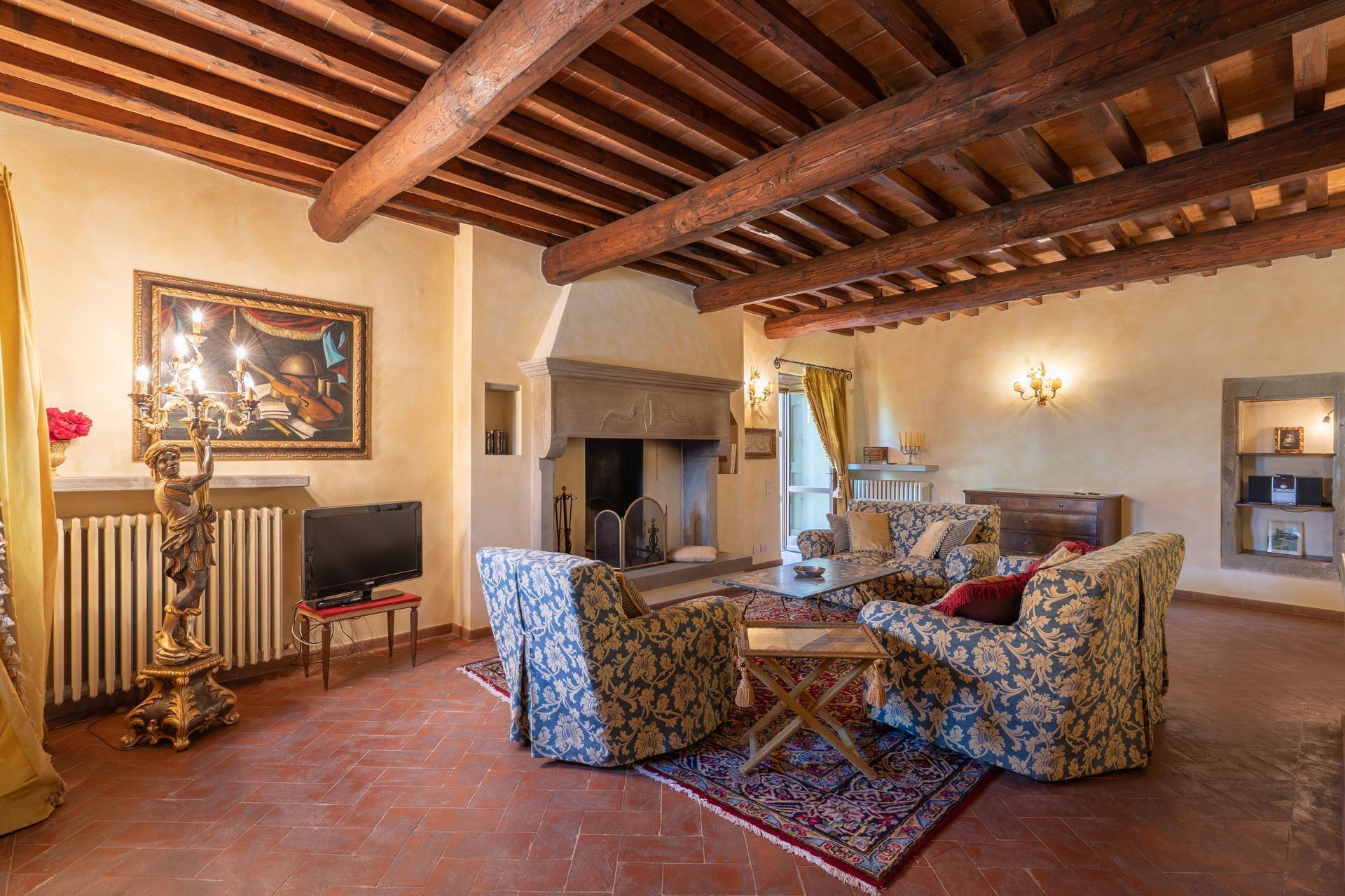 A cozy living room with a fireplace, patterned sofas, a wooden ceiling, and a television.