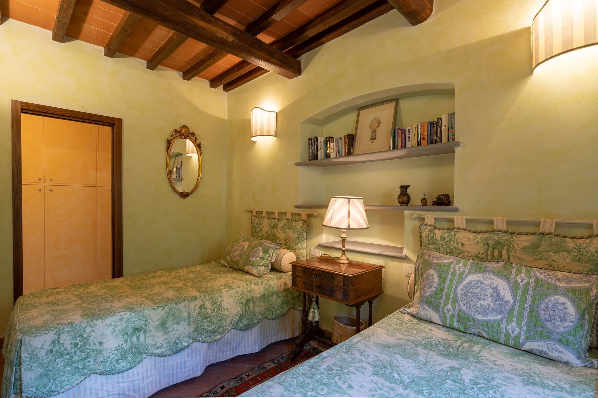 Une chambre avec deux lits simples, une armoire en bois, un miroir, une table de chevet avec une lampe et une bibliothèque contenant des livres et des objets décoratifs.