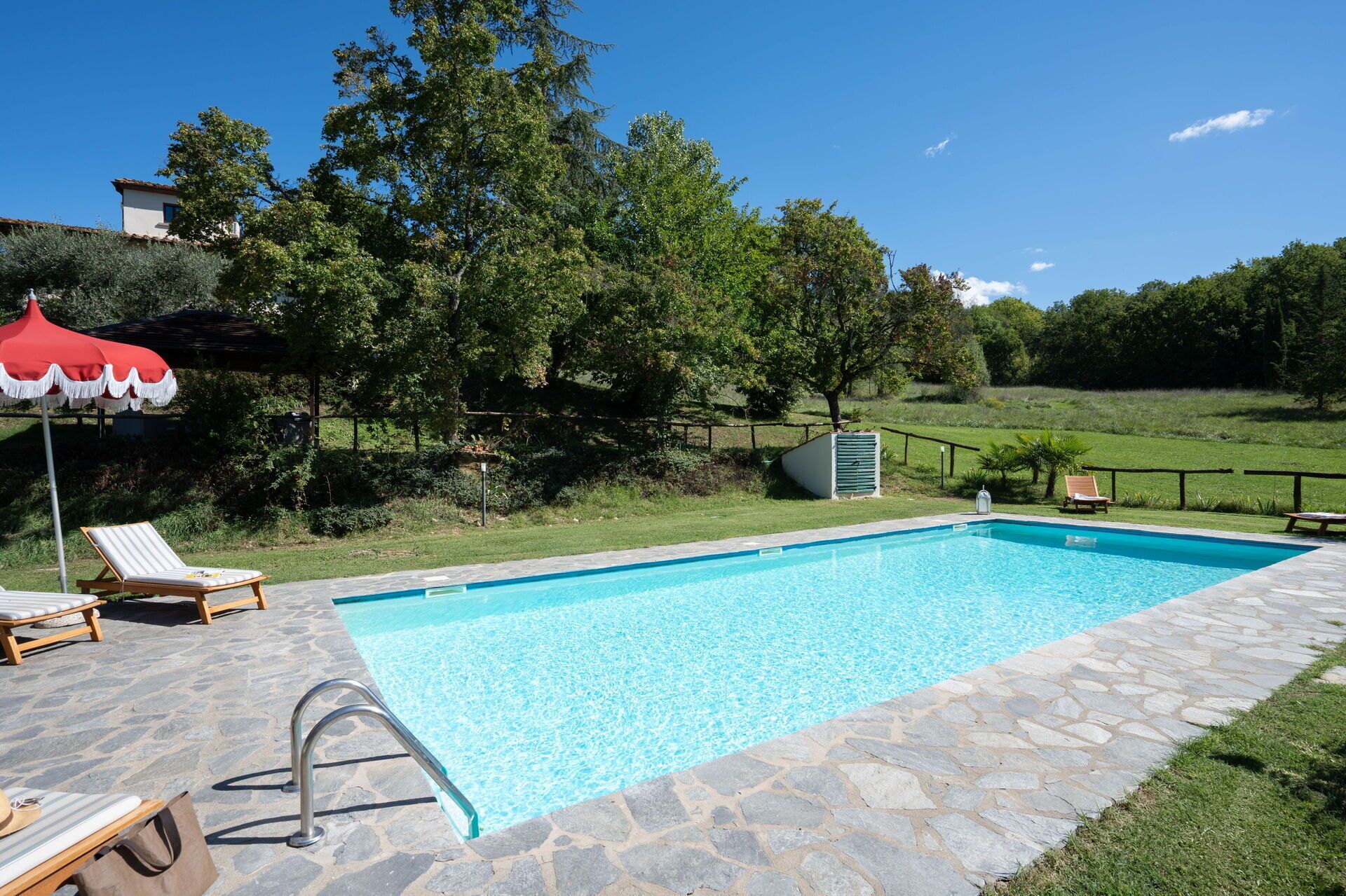 A rectangular swimming pool with stone tiles, surrounded by a stone deck. There are lounge chairs and a red umbrella.