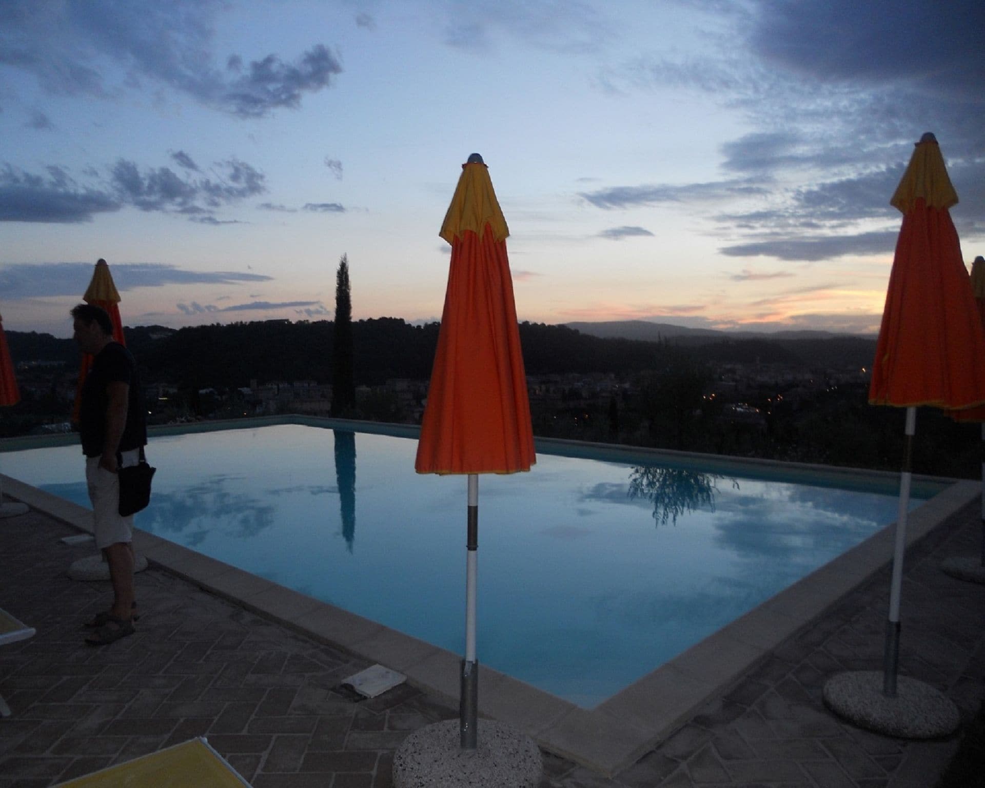 A swimming pool with three umbrellas, a person standing by the pool, and a cityscape in the background.
