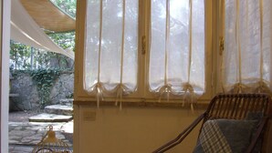 Une pièce avec un fauteuil en osier, une petite table sur laquelle est posé un vase décoratif, et une vue sur un jardin, visible à travers une fenêtre ornée de rideaux légers.