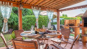 Een terras buiten met een bakstenen barbecue, houten stoelen en een eettafel.
