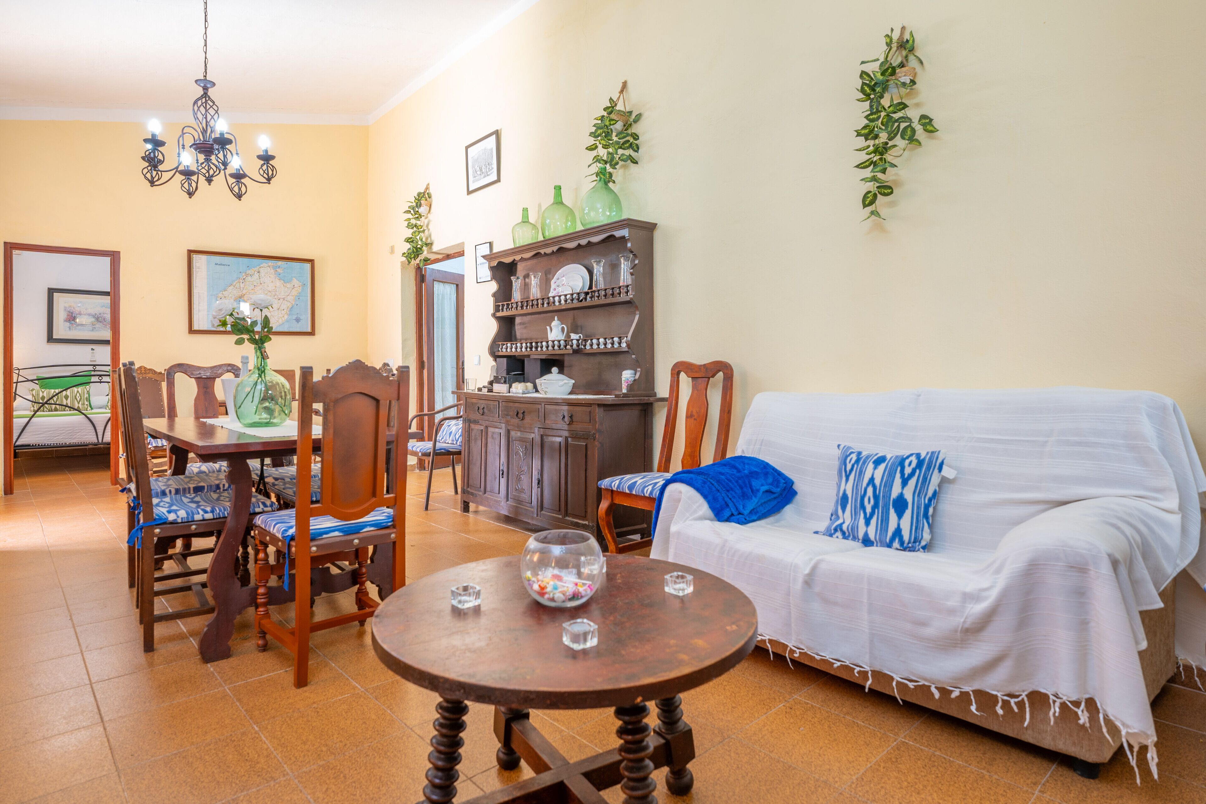 Un comedor con mesa de madera, sillas y una araña. Hay un sofá con un cojín azul y una manta blanca. También se observa un mueble con vajilla y un mapa en la pared.