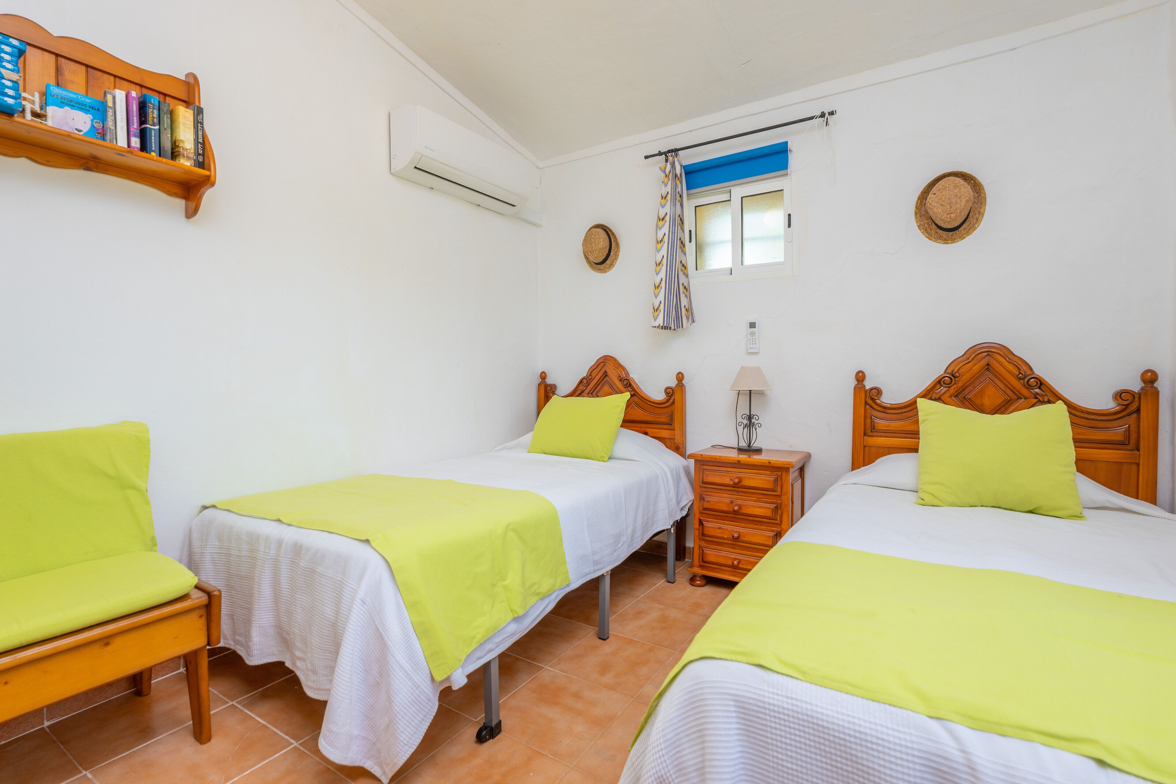 A room with two single beds, each with a wooden headboard, a bedside table, and a chair. There is a shelf with books and a wall-mounted air conditioner.