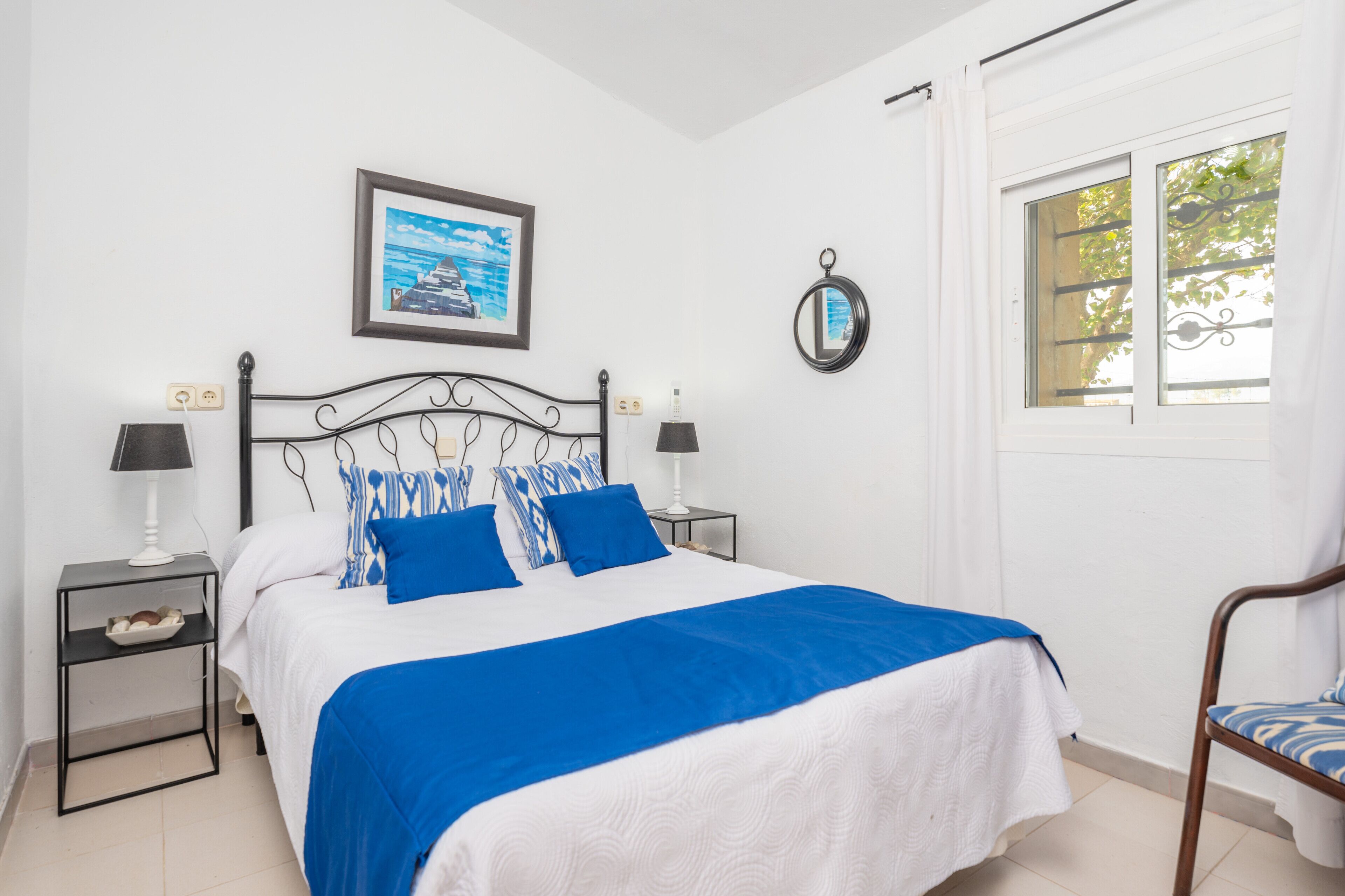 A bedroom with a bed, two bedside tables, a chair, a window, and a framed picture on the wall.