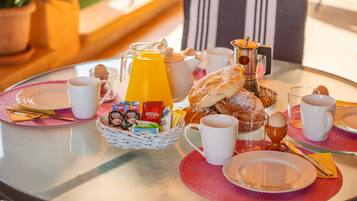 A breakfast table with a glass pitcher of orange juice, a coffee pot, pastries, and a basket of assorted snacks.