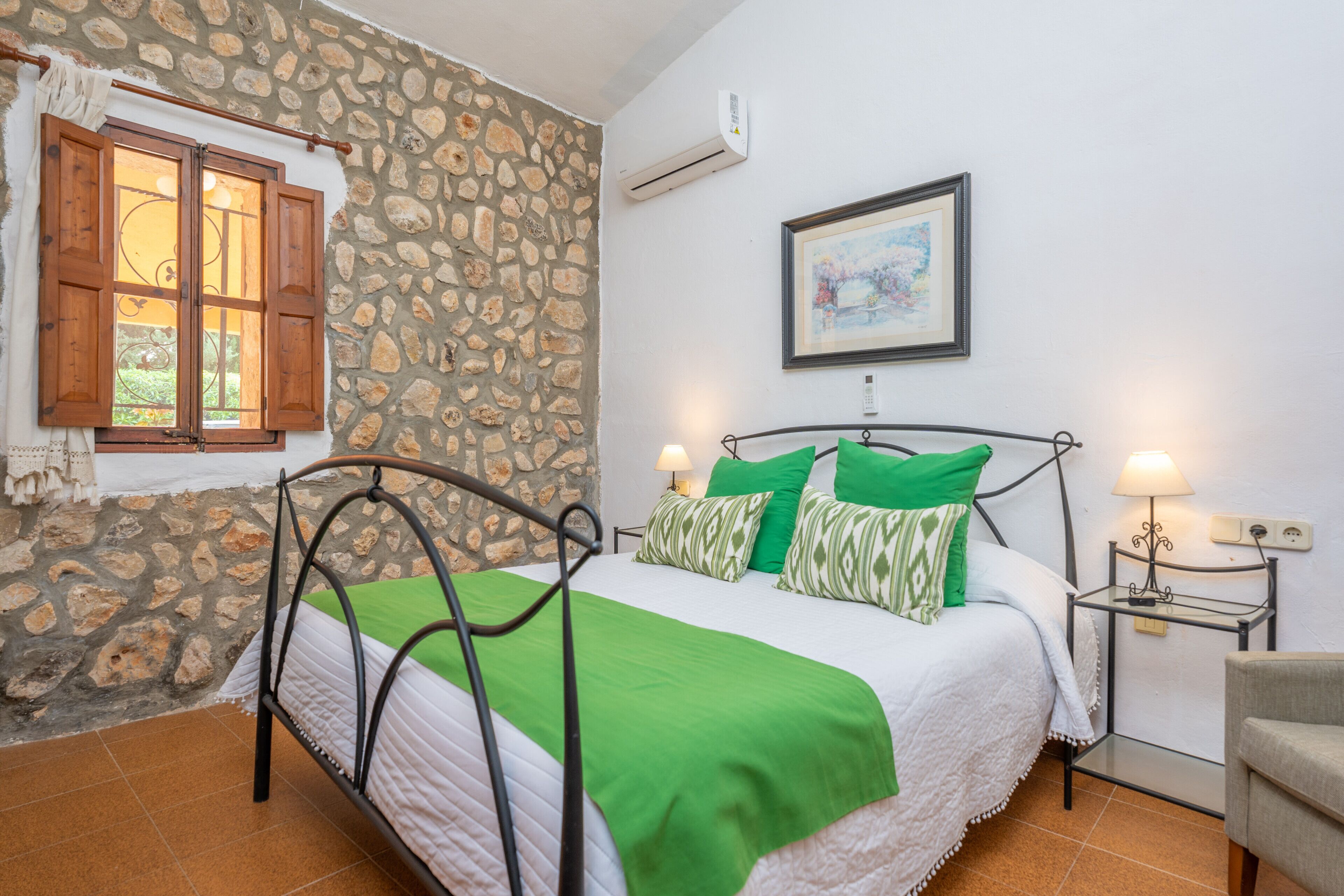 Quarto com parede de pedra, cama com roupa de cama verde, janela de madeira e um quadro pendurado na parede.