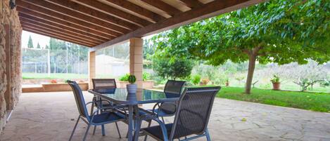 A covered patio with a table and chairs, stone flooring, and a view of a garden with trees and plants.