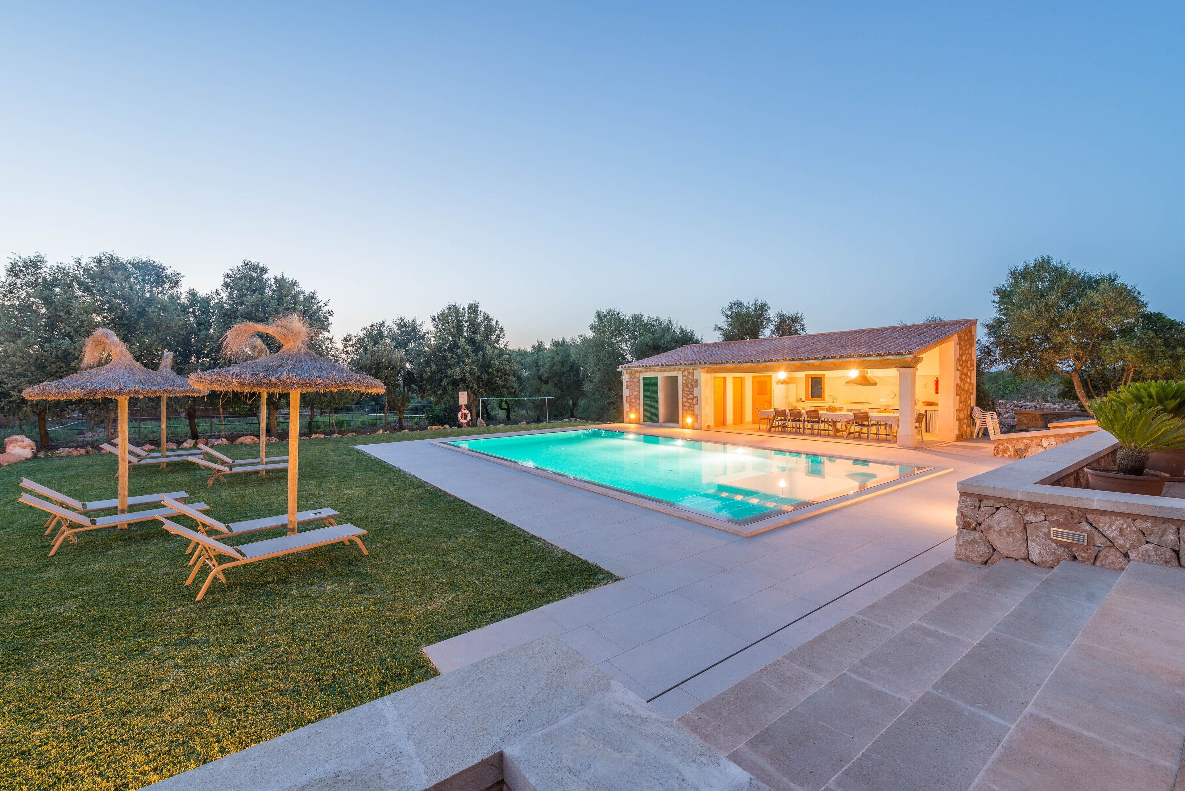 A swimming pool with lounge chairs and a covered seating area, surrounded by a stone patio and a house with a tiled roof.