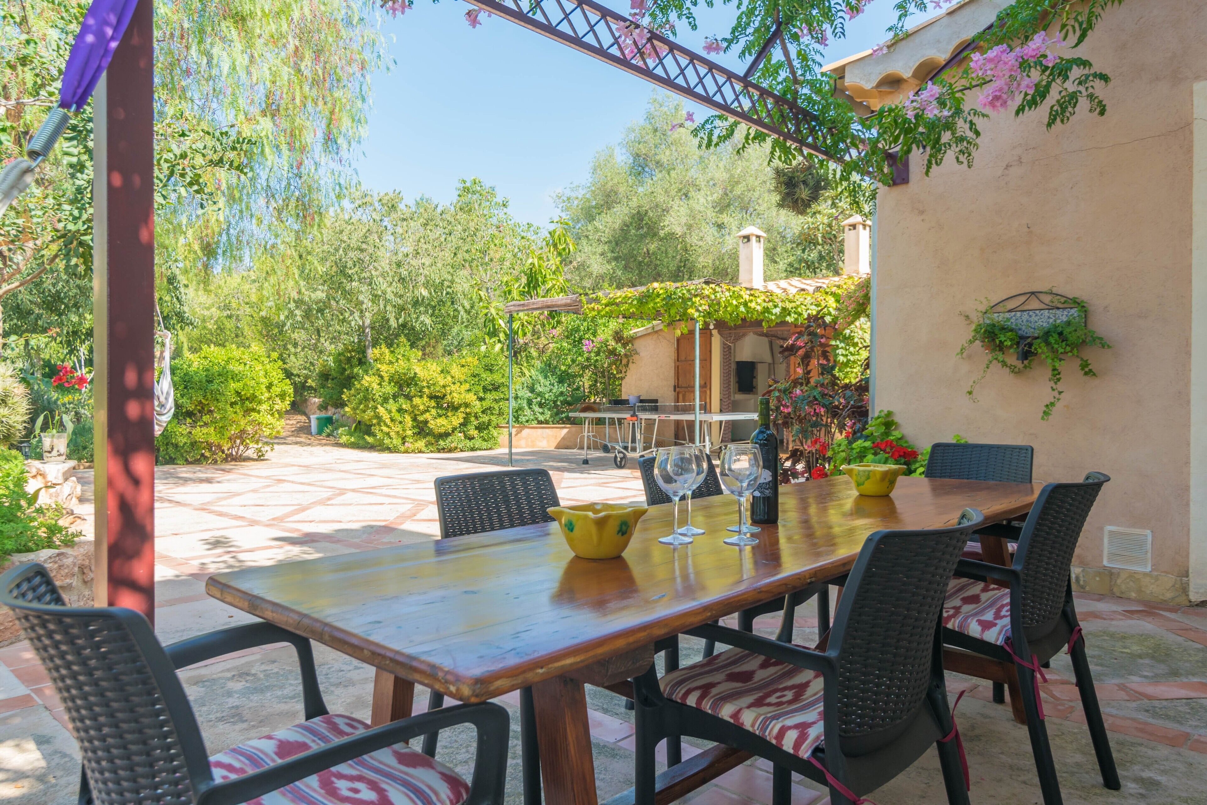 Un patio al aire libre con una mesa y sillas de madera, rodeado de follaje y una pérgola.