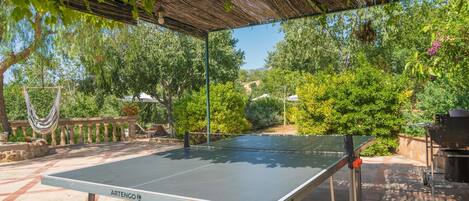 Une table de ping-pong installée à l’extérieur, sous une pergola, avec vue sur un jardin et des arbres.