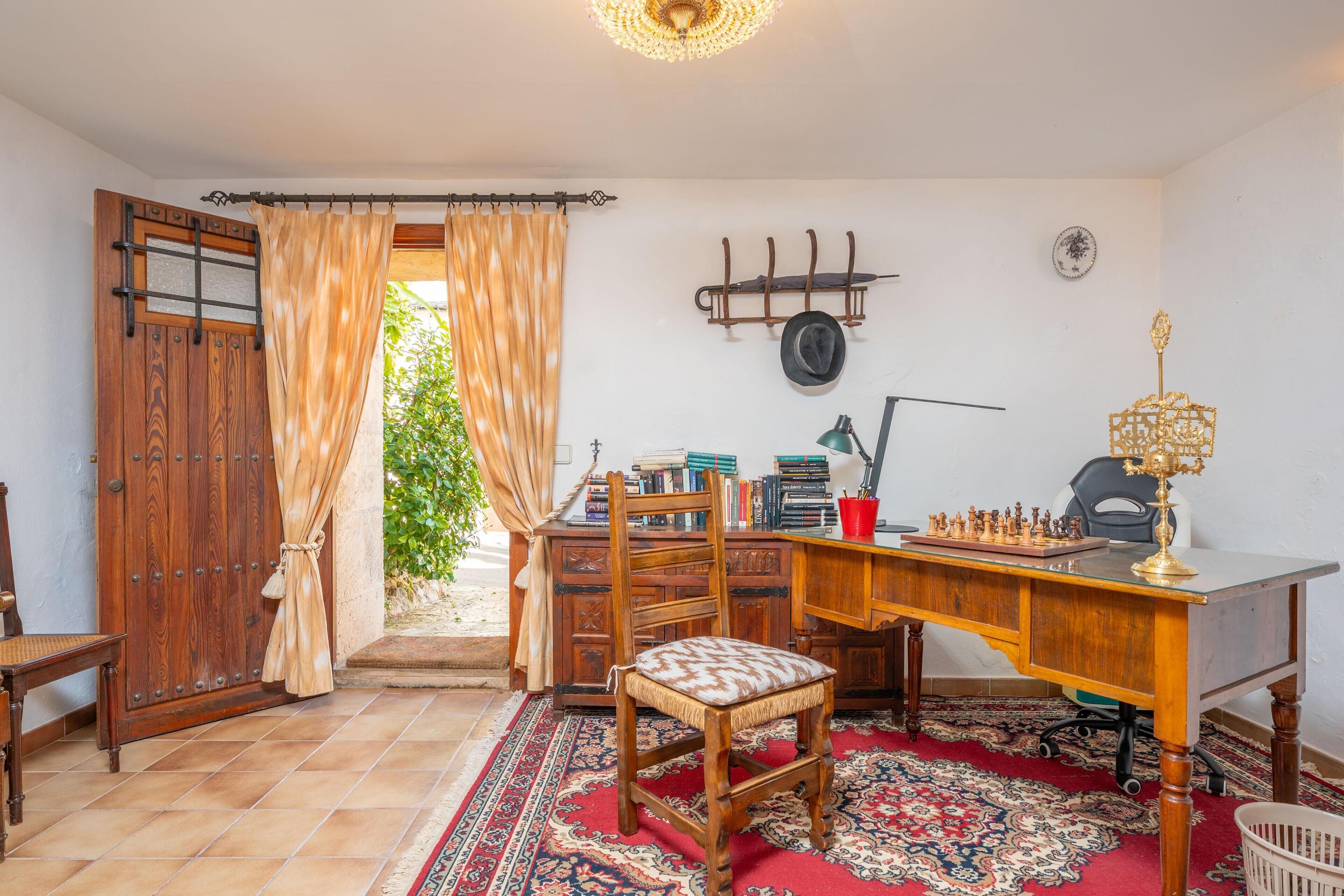 A room with a wooden desk, a chair, a chessboard, a lamp, and a red patterned rug.