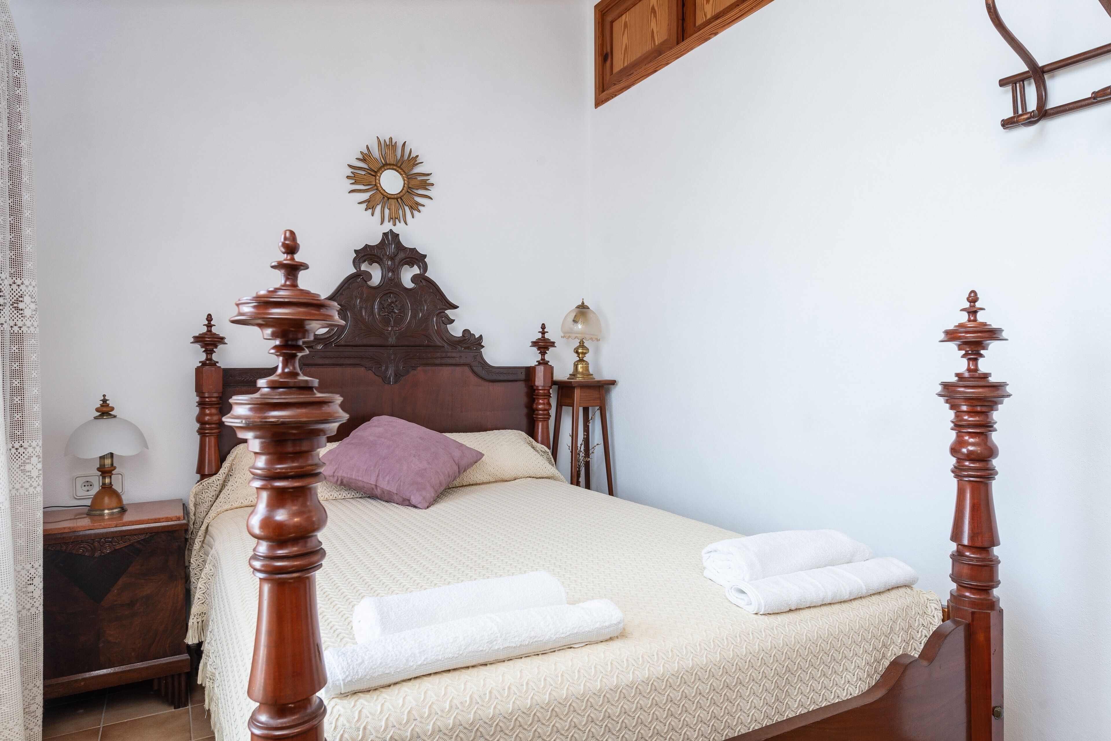 A neatly made bed with a wooden headboard, a bedside table with a lamp, and a decorative sun-shaped mirror on the wall.