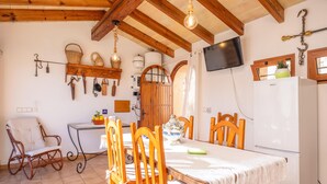 A rustic kitchen with wooden beams, a dining table with chairs, a refrigerator, and a television mounted on the wall.