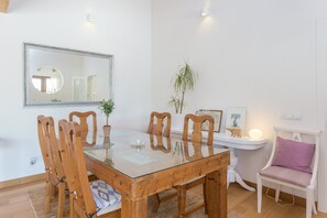 Ein Essbereich mit einem Glastisch, Holzstühlen, einem weißen Stuhl mit violettem Sitzkissen, einem Spiegel an der Wand und einer kleinen Pflanze auf dem Tisch.