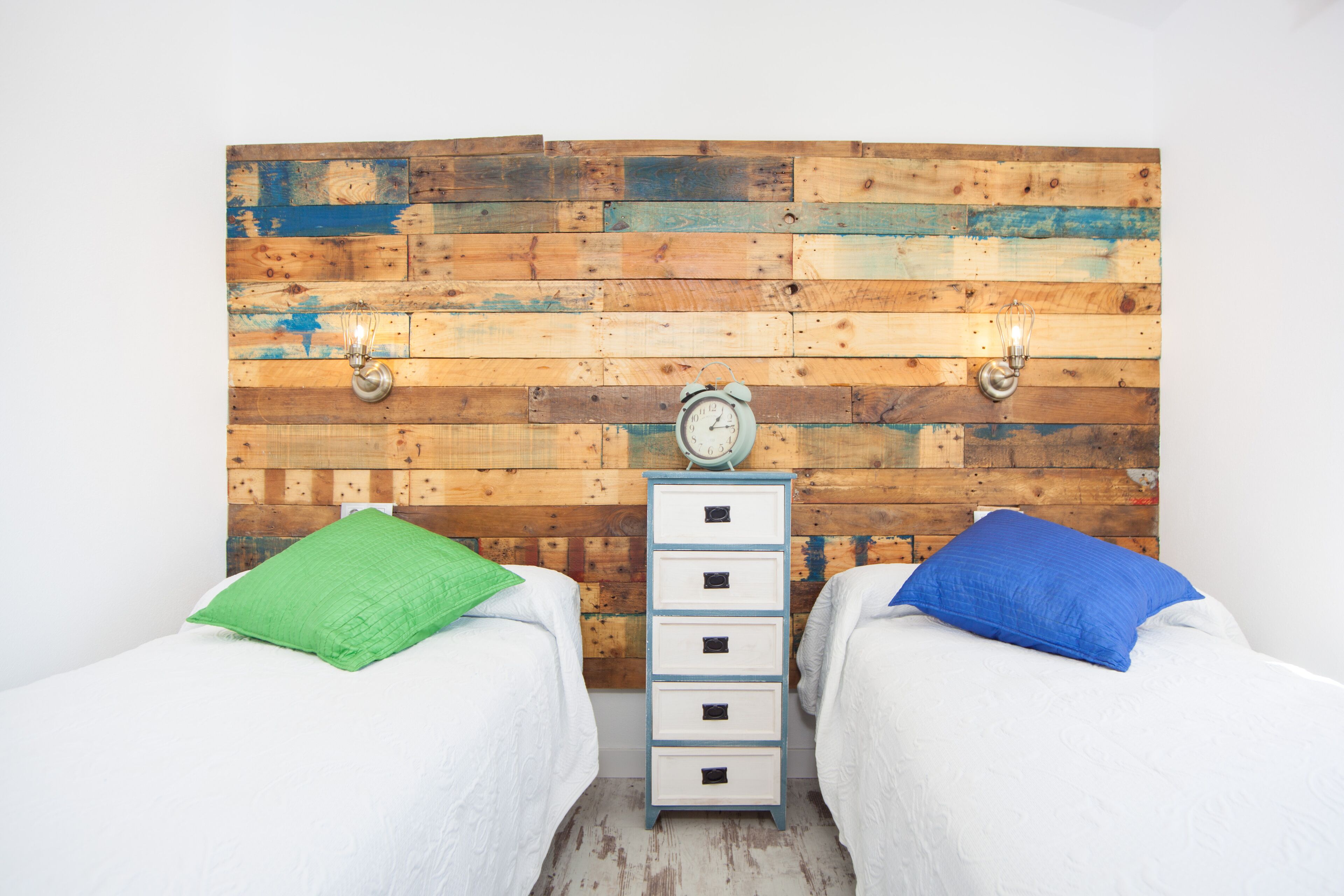 Une chambre avec deux lits, chacun équipé d’un oreiller, d’une commode avec des tiroirs et d’une tête de lit en bois agrémentée d’une horloge et d’appliques murales.
