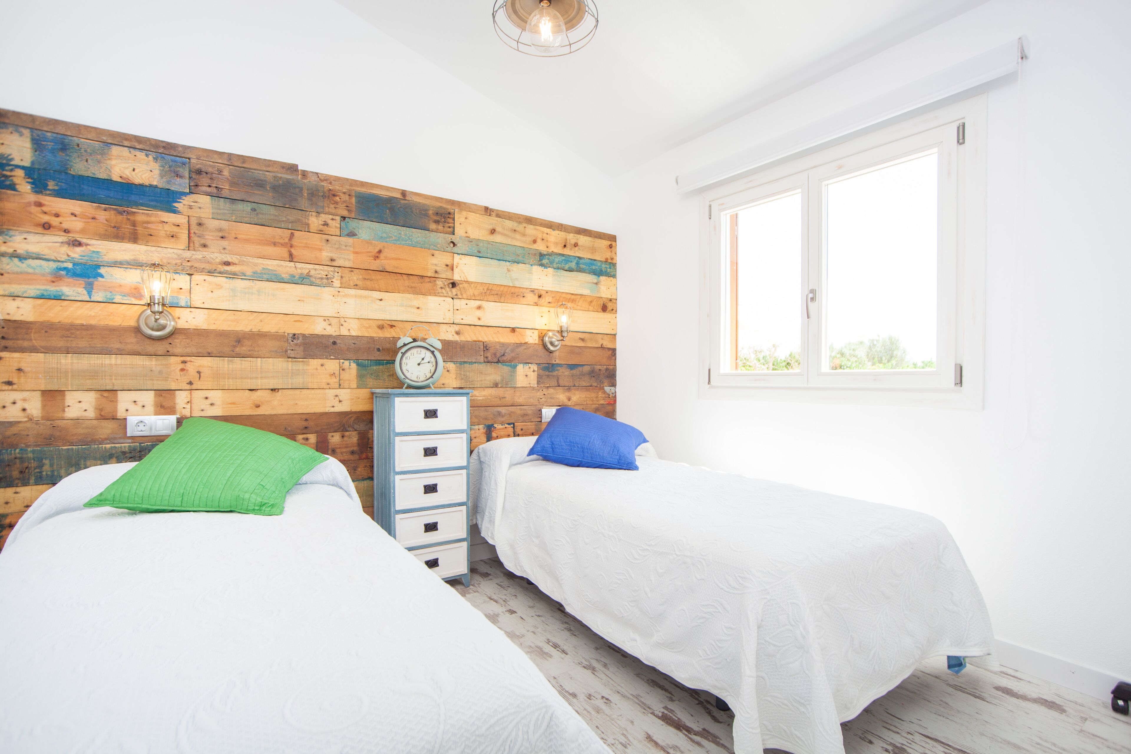 A bedroom with two beds, a wooden wall panel, a clock, a dresser, and a window.