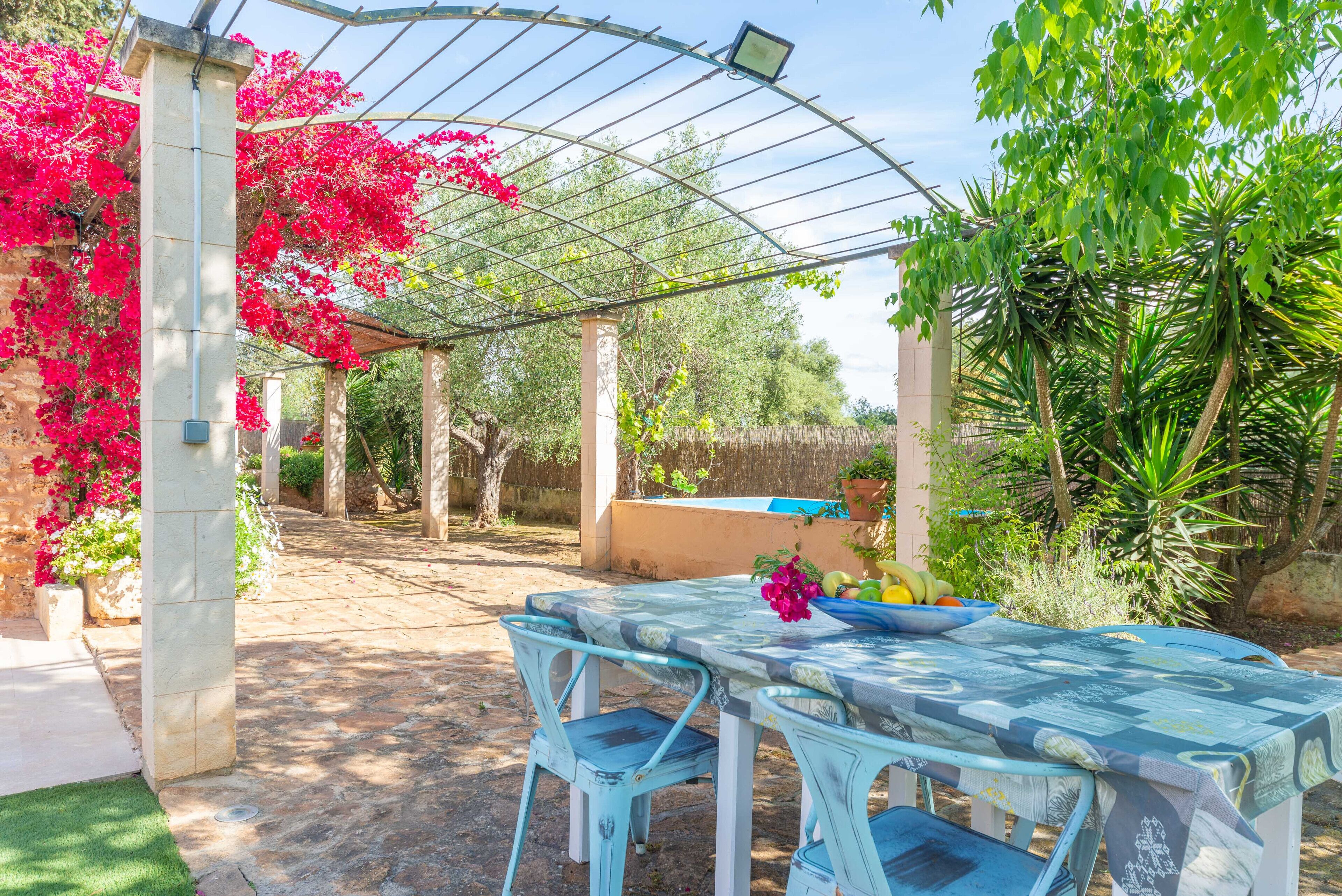 A covered patio area with a table and chairs, surrounded by greenery and a pergola structure.