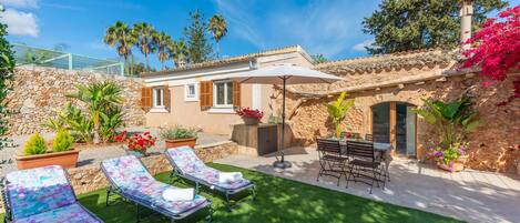A stone house with a patio, two sun loungers, a table with chairs, and a large umbrella.