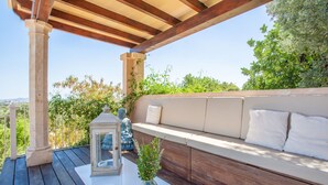 Ein Sitzbereich im Freien mit einer Holnpergola, einer gepolsterten Bank mit weißen Kissen und einer Glaskaraffe mit Grünpflanzen.