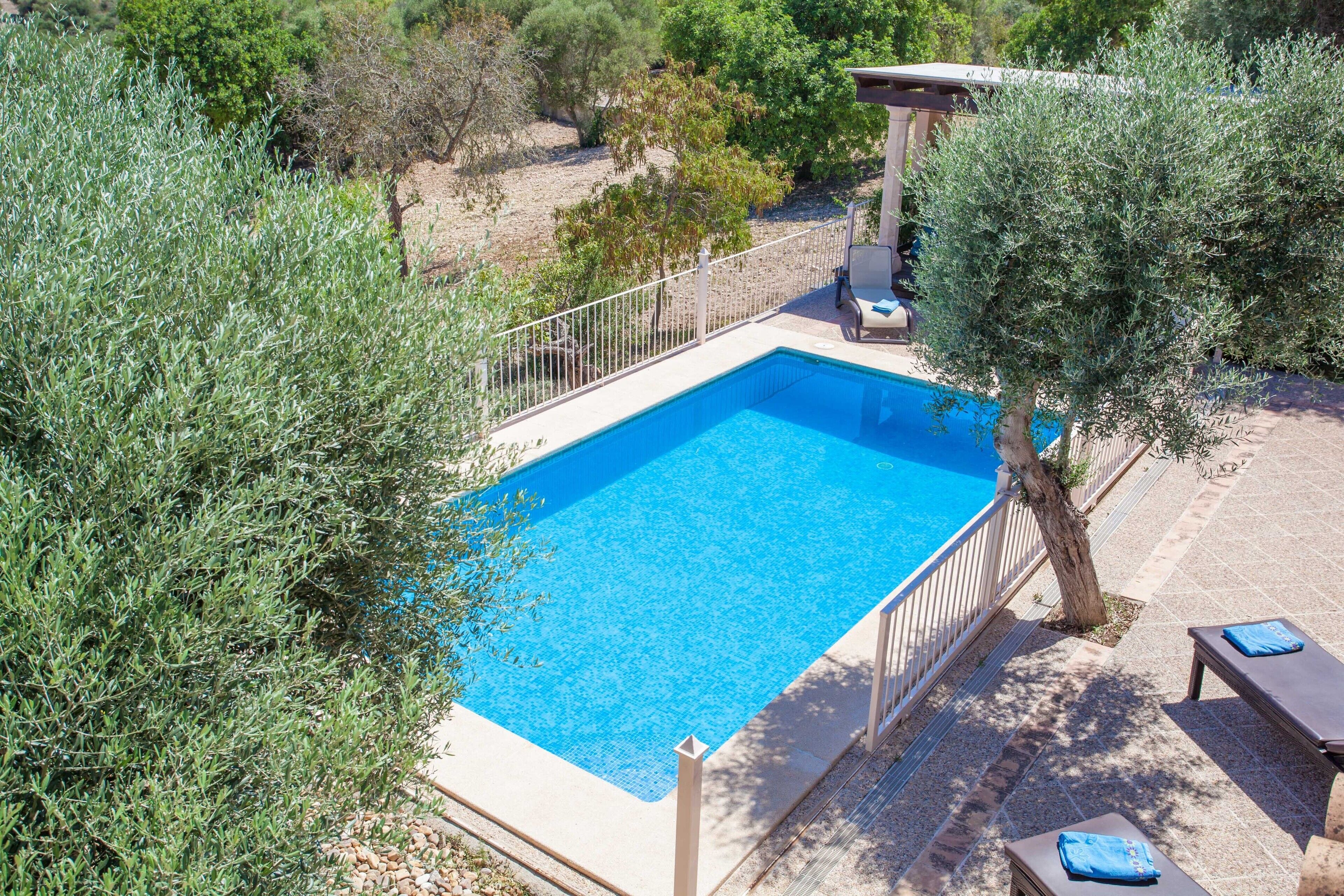 Una piscina rectangular con una escalera blanca, rodeada por una valla blanca. Se observan dos sillones de jardín con cojines azules y una estructura de pérgola al fondo.