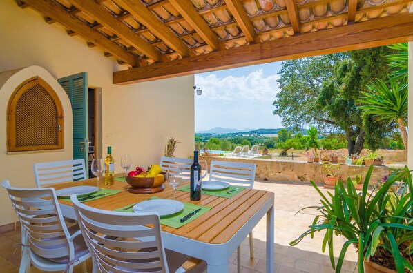 An outdoor dining area with a wooden table and white chairs, a bottle of wine, and a bowl of fruit.